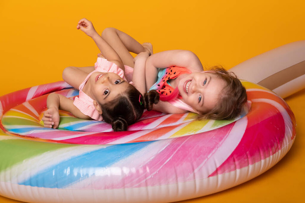 dos niña en traje de baño que miente que se divierte en la piruleta inflable colorida del colchón en fondo amarillo - Foto, imagen