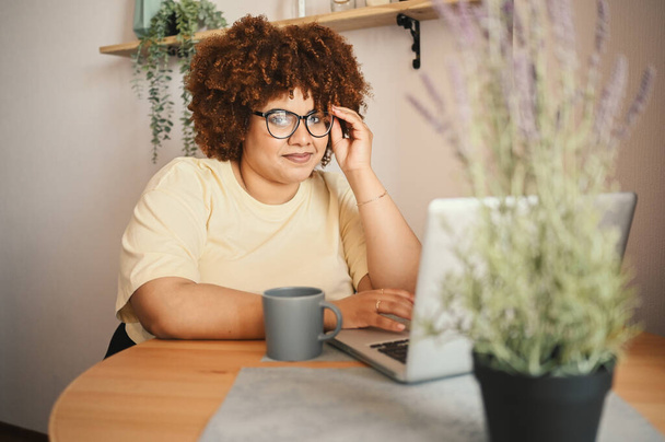 Vonzó boldog elegáns plusz méret Afrikai fekete diák afro haj szemüvegben tanul online dolgozik laptop számítógép otthoni irodai munkaterület. Sokszínűség. Távmunka, távoktatás. - Fotó, kép