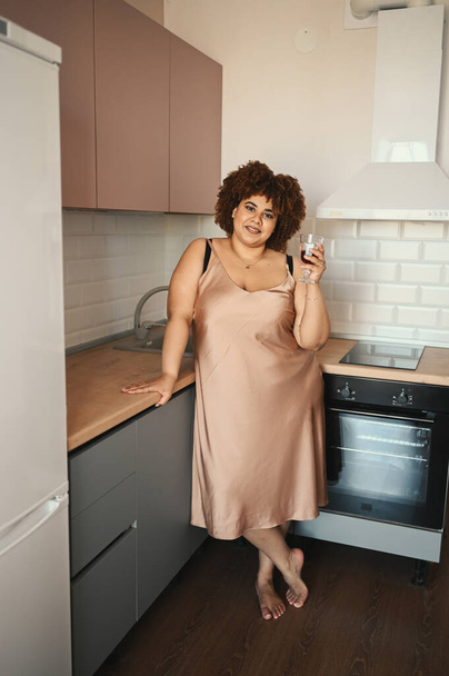 Beautiful curvy plus size African black woman afro hair drinking red wine on modern scandinavian style kitchen interior design. Body imperfection, body acceptance, body positive and diversity concept - 写真・画像