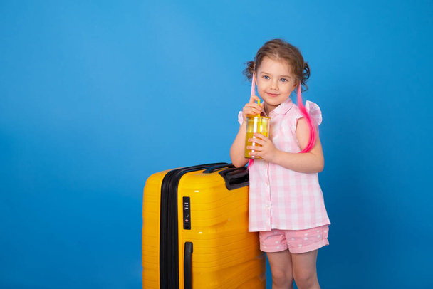 Zabawna dziewczynka w różowych ubraniach trzyma szklankę soku pomarańczowego z żółtą walizką na niebieskim tle. - Zdjęcie, obraz