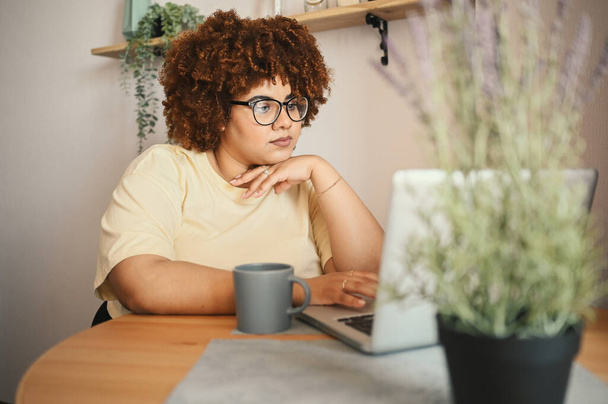 Atrakcyjny szczęśliwy stylowy plus size Afrykańska czarna studentka afro włosy w okularach studiuje online pracując na laptopie w biurze domu przestrzeni roboczej. Różnorodność. Praca zdalna, kształcenie na odległość. - Zdjęcie, obraz