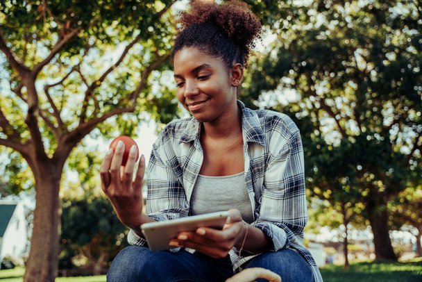 Mixed race female teen researching for project on digital tablet while holding fresh tomato  - Фото, изображение