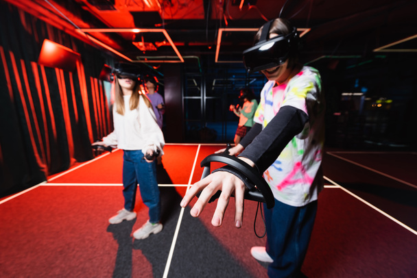 VR kulaklıklı neşeli çocuk oyun odasında arkadaşlarıyla el kol hareketi yapıyor. - Fotoğraf, Görsel