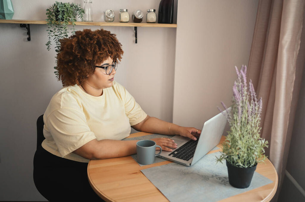 Attractive happy stylish plus size African black woman student afro hair in glasses studying online working on laptop computer at home office workspace. Diversity. Remote work, distance education. - Photo, Image