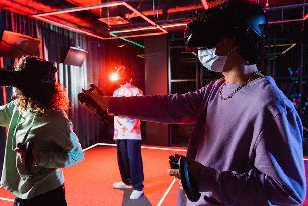 multi-etnische tiener vrienden in medische maskers spelen vr spel in headsets - Foto, afbeelding