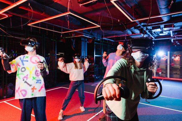 multiethnic friends in medical masks and vr headsets having fun in play room - Photo, Image