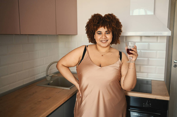Gyönyörű görbe plusz méret Afrikai fekete nő afro haj ivás vörösbor a modern skandináv stílusú konyha belsőépítészet. A test tökéletlensége, a test elfogadása, a test pozitív és sokszínű koncepciója - Fotó, kép