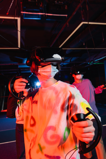 мальчик-подросток в медицинской маске игры вблизи размытых друзей в виртуальной игровой зоне - Фото, изображение