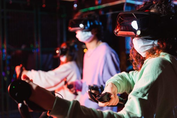 african american girl in medical mask and vr headset gaming near blurred friends - Photo, Image