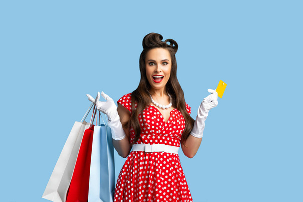 Привлекательная молодая женщина пинап в платье в стиле ретро с подарочными пакетами и кредиткой на фоне синей студии - Фото, изображение