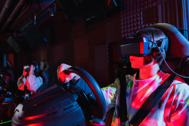 gamer in vr headset on car racing simulator near blurred friend - Foto, afbeelding