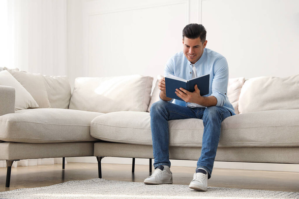 Man with book sitting on comfortable sofa in living room - Foto, Imagen