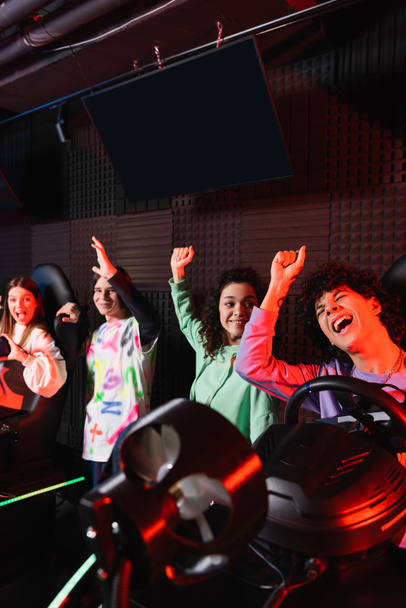 excited multiethnic friends showing rejoice gesture near racing simulators - Foto, afbeelding
