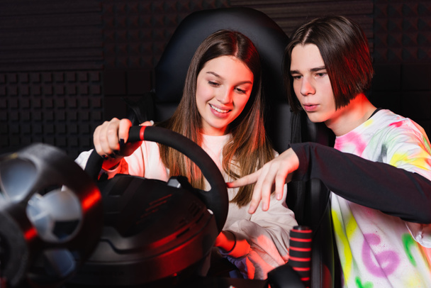 adolescent garçon pointant au volant de simulateur de voiture près ami souriant - Photo, image