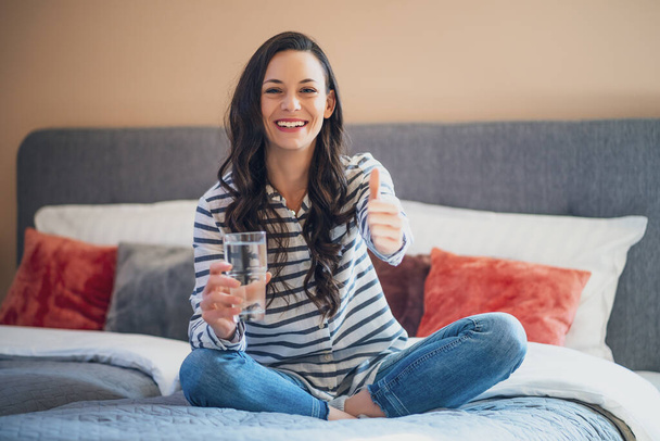 Happy young woman sitting on bed in her home, looking at camera and holding glass of water. - Photo, image