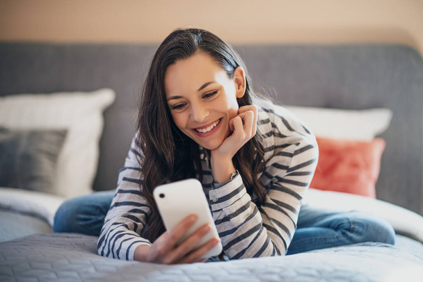 幸せな若い女性は彼女の家のベッドの上に横たわって、スマートフォンを使用して. - 写真・画像