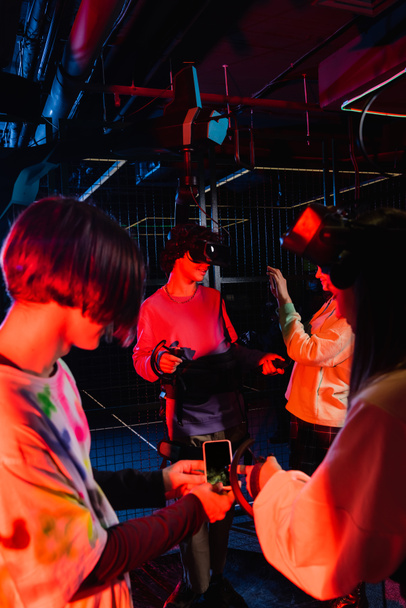 adolescente sosteniendo teléfono inteligente con pantalla en blanco cerca de amigos en la zona de juego vr - Foto, imagen