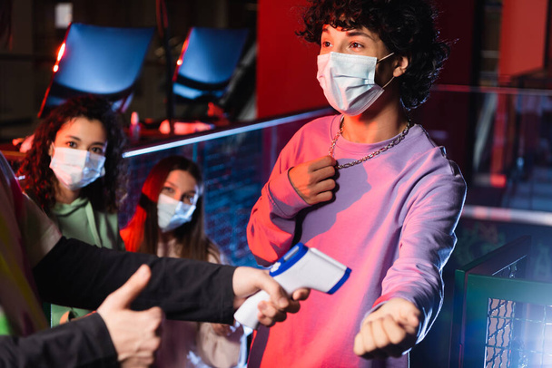 controlador de zona de jogo mostrando polegar perto de adolescentes inter-raciais em máscaras médicas - Foto, Imagem