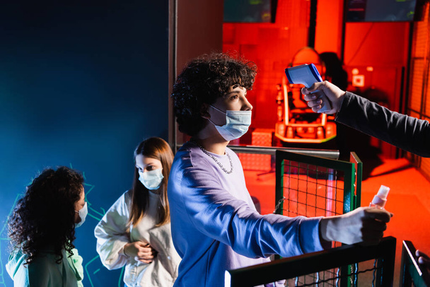 Teenager-Gamer in medizinischer Maske hält Händedesinfektionsmittel in der Nähe von Controller mit Pyrometer und interrassischen Freunden - Foto, Bild