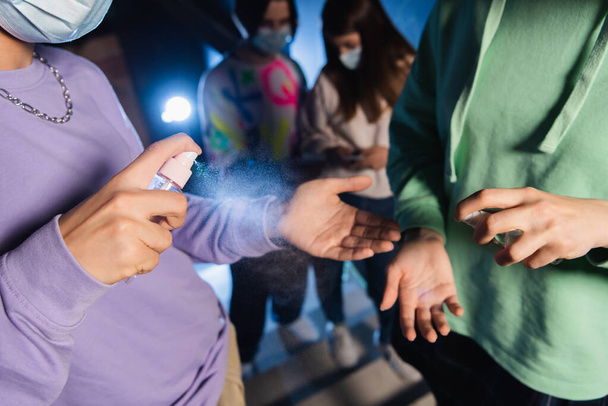διαφυλετικοί παίκτες που εφαρμόζουν απολυμαντικό χεριών κοντά σε θολή φίλους σε ιατρικές μάσκες - Φωτογραφία, εικόνα
