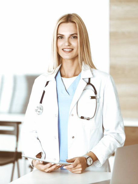 Woman-doctor at work in clinic excited and happy of her profession. Blond female physician is smiling while using tablet computer. Medicine concept - Foto, Bild