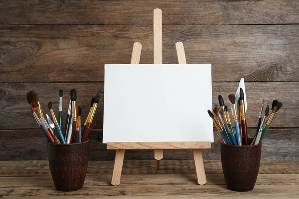 Easel with blank canvas and brushes on wooden table - Foto, Bild