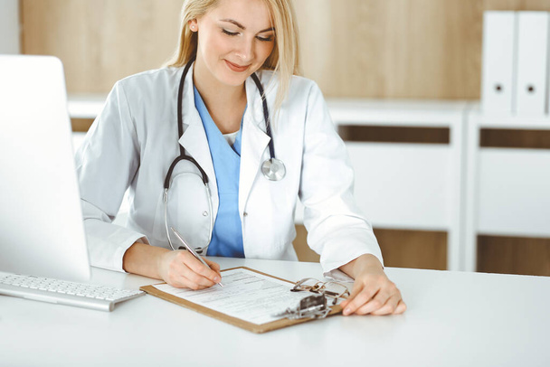 Woman-doctor at work while sitting at the desk in hospital or clinic. Blonde cheerful physician filling up medication history record form - Photo, Image