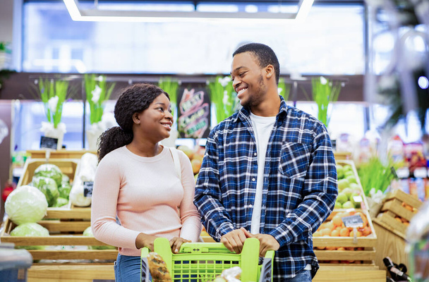 Portrét usmívající se mladé černošky s nákupním vozíkem při pohledu na sebe v zeleninové části obchodu - Fotografie, Obrázek