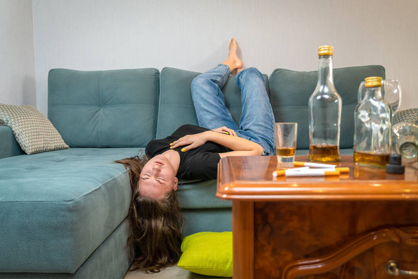 Пьяная молодая темноволосая женщина спит на диване после вечеринки, пустые бутылки на столе - Фото, изображение