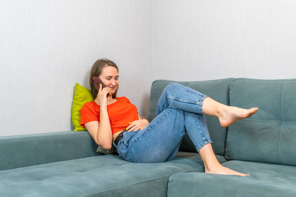 Jeune belle femme brune en jean bleu et une chemise orange est allongée sur le canapé et parle au téléphone - Photo, image