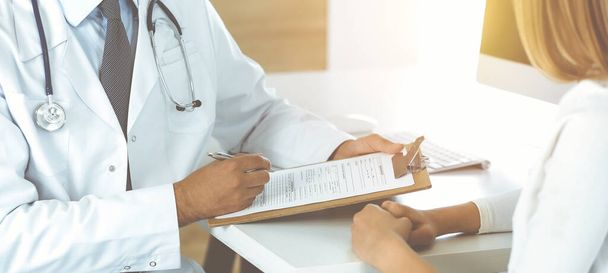 Doctor and patient discussing medical exam results while sitting at the desk in sunny clinic, close-up. Male physician using clipboard. - Foto, Imagen