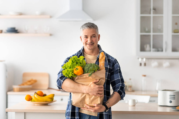 Happy mature man holding paper bag full of groceries from the supermarket, standing in kitchen interior, copy space. - Foto, imagen
