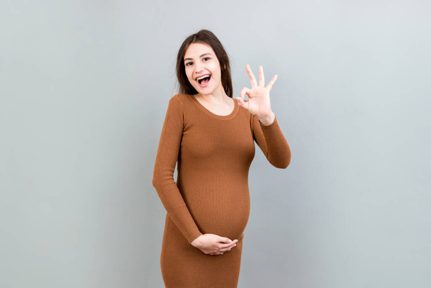 положительная беременная женщина показывает нормальный жест на животе на цветном фоне. Легкая и счастливая беременность Копирование пространства. - Фото, изображение