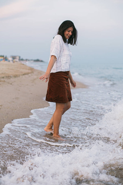 Happy carefree hipster woman walking barefoot in cold sea waves on  sandy beach, enjoying tranquil evening. Casual young female relaxing and having fun on seashore at resort. Mindfulness - Photo, Image