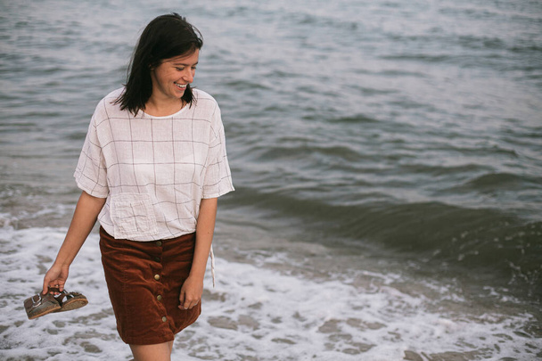 Szczęśliwa uśmiechnięta hipsterka spacerująca boso po piaszczystej plaży i falach morskich wieczorem, ciesząca się spokojną chwilą. Zwykła młoda samica z japonkami w ręku relaksująca się na brzegu morza - Zdjęcie, obraz