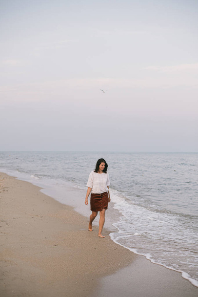 Carefree hipster woman walking barefoot on sandy beach with sea waves in the evening, enjoying tranquil moment. Casual young female  relaxing on seashore at resort. Mindfulness - Fotó, kép