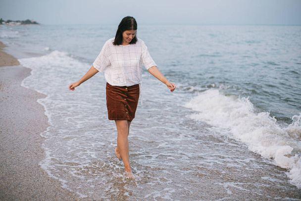 Беззаботная хипстерша гуляет босиком по песчаному пляжу с морскими волнами вечером, наслаждаясь спокойным моментом. Повседневная молодая женщина отдыхает на берегу моря на курорте. Памятование - Фото, изображение