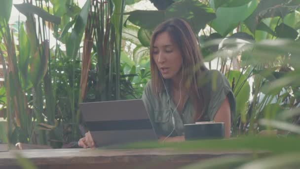 Poitrine-up de femme caucasienne aux cheveux longs portant un casque filaire, assise à la table à l'extérieur parmi de grandes plantes vertes lors d'une chaude journée d'été, en utilisant un ordinateur portable pour la conversation en ligne - Séquence, vidéo