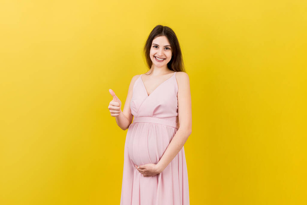 Счастливая беременная женщина показывает большой палец вверх крутой жест над ее ребенка шишка на цветном фоне. Счастливого материнства. Копирование пространства. - Фото, изображение