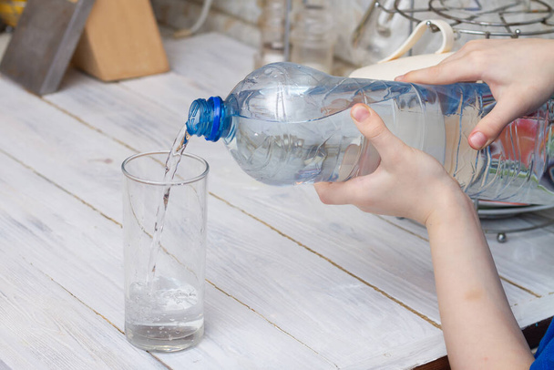 Pour water into a glass glass from a plastic bottle in the kitchen at home. - Φωτογραφία, εικόνα