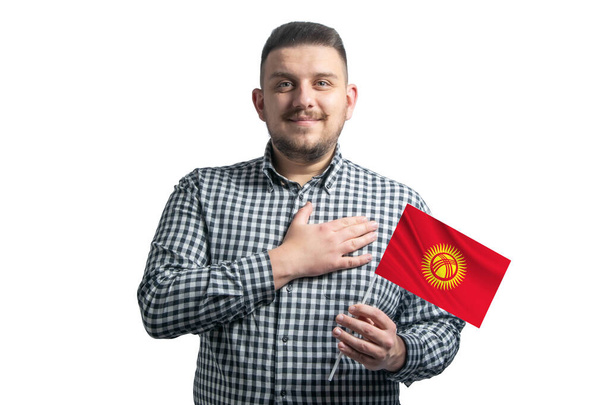 Hombre blanco sosteniendo una bandera de Kirguistán y sostiene su mano sobre su corazón aislado sobre un fondo blanco Con amor a Kirguistán. - Foto, imagen