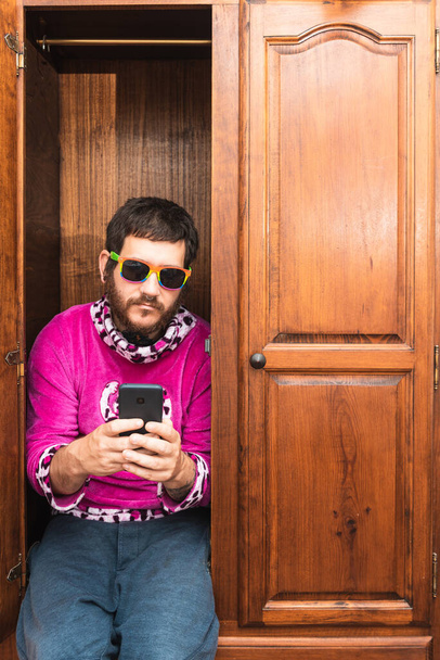 Homosexuální muž s piercing v nose a duhovými brýlemi s vážným gestem sedí ve skříni a používá svůj smartphone. Vylez ze skříně. Komunita Igbt. - Fotografie, Obrázek