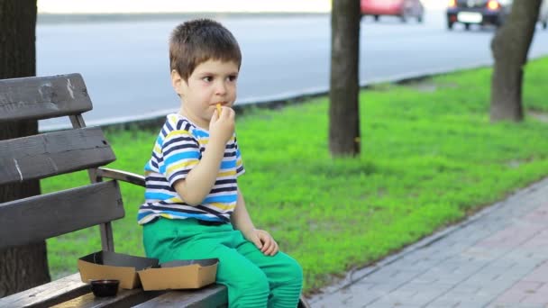 Un bambino piccolo mangia patatine fritte su una strada della città, seduto su una panchina. Getta il cibo a terra. Fast food per bambini - Filmati, video