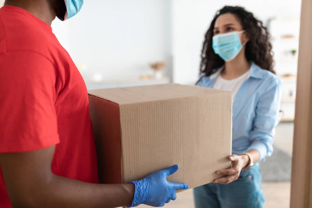zwart levering man in medisch masker geven doos naar vrouw - Foto, afbeelding