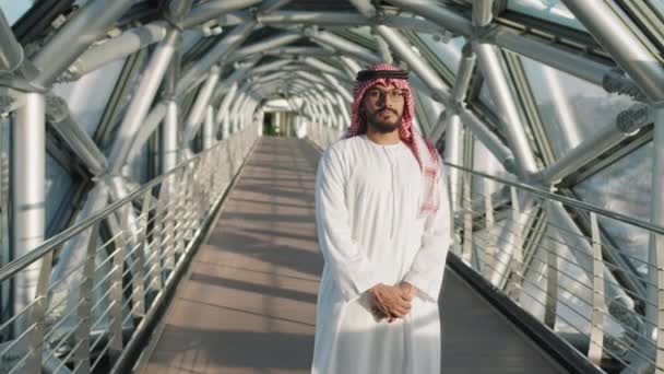 Antenne Medium Slowmo Porträt eines arabischen Mannes in traditioneller Kleidung, der in die Kamera blickt, die auf dem gläsernen Gehweg eines modernen Bürogebäudes der gehobenen Klasse steht - Filmmaterial, Video