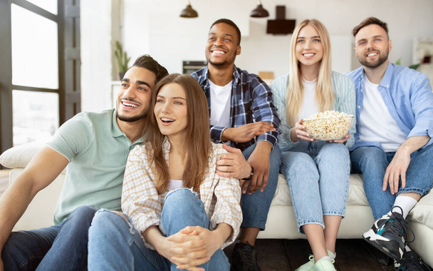 Группа веселых молодых друзей, наслаждающихся кино дома, кушающих попкорн и улыбающихся - Фото, изображение