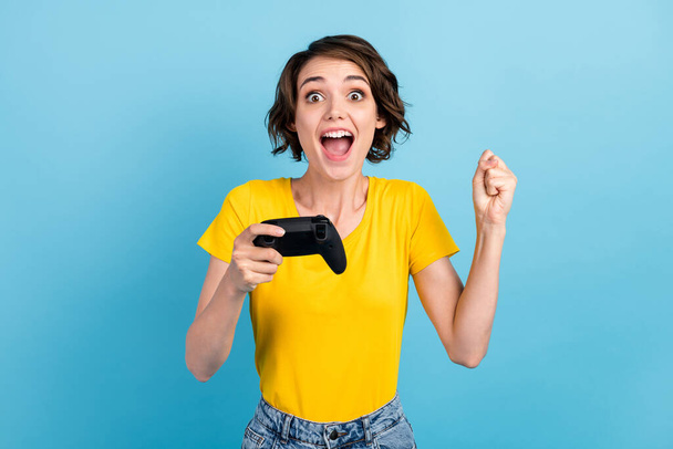 Photo of excited girl hold controller open mouth raise fist wear yellow t-shirt isolated blue color background - Foto, afbeelding