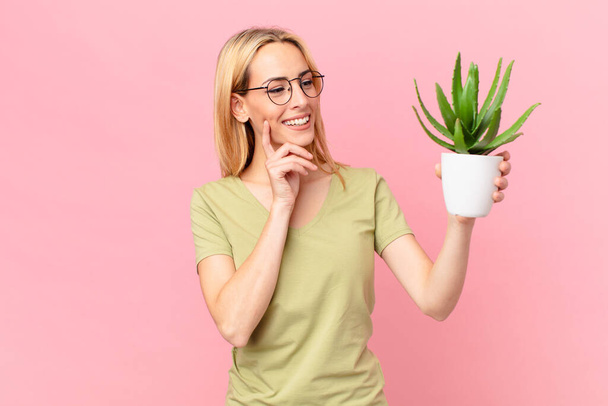 young blonde woman smiling happily and daydreaming or doubting and holding a cactus - Foto, Imagem