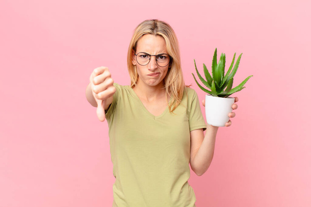 young blonde woman feeling cross,showing thumbs down and holding a cactus - Fotó, kép