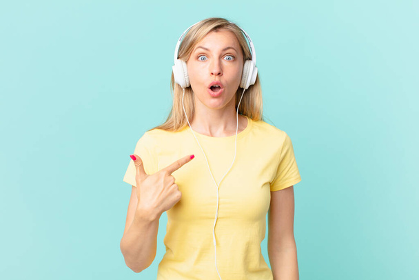 young blonde woman looking shocked and surprised with mouth wide open, pointing to self and listening music. - Foto, Imagen